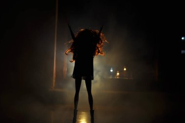 Hafif arka planda bir kadının silueti. Geceleri uzun saçlı, arka planda şehir ışıkları olan yalnız bir bebeğin resmi. Seçici odak