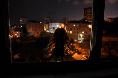 Hafif arka planda bir kadının silueti. Geceleri uzun saçlı, arka planda şehir ışıkları olan yalnız bir bebeğin resmi. Seçici odak