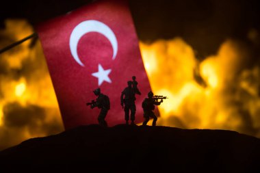 Yanan karanlık arka planda küçük bir hindi bayrağı. Savaş krizi kavramı ve uluslar arasındaki siyasi çatışmalar. Türk bayrağına karşı silahlı asker silueti. Seçici odak
