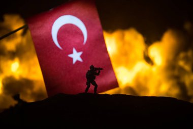 Yanan karanlık arka planda küçük bir hindi bayrağı. Savaş krizi kavramı ve uluslar arasındaki siyasi çatışmalar. Türk bayrağına karşı silahlı asker silueti. Seçici odak