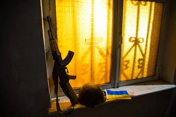 Conceptual photo of war between Russia and Ukraine. Ukrainian flag, helmet and gun on windowsill at night. Old creepy room with window. Explosion outside.