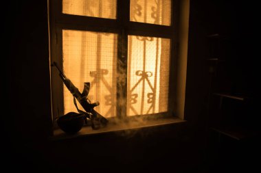 Rusya ve Ukrayna arasındaki savaşın kavramsal fotoğrafı. Rus silahı ve miğferi gece pencere eşiğinde. Pencereli eski ürkütücü oda. Dışarıdaki patlama..