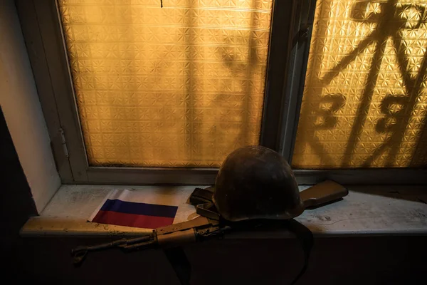 Conceptual photo of war between Russia and Ukraine. Russian flag military helmet and weapon on windowsill at night. Old creepy room with window. Explosion outside.