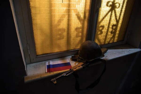 Conceptual photo of war between Russia and Ukraine. Russian flag military helmet and weapon on windowsill at night. Old creepy room with window. Explosion outside.