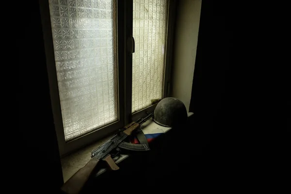 Conceptual photo of war between Russia and Ukraine. Russian flag military helmet and weapon on windowsill at night. Old creepy room with window. Explosion outside.