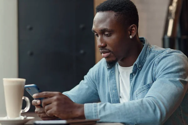 Empresario negro concentrado utiliza teléfono inteligente mientras está sentado en la cafetería con café cerca de la ventana. — Foto de Stock