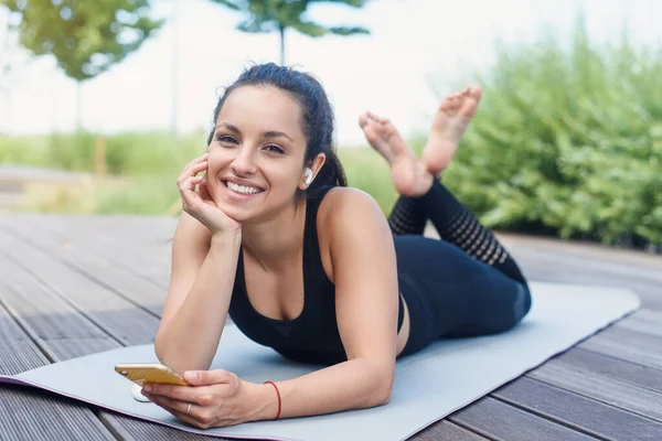 Woman lying with phone on yoga mat after exercising, relaxing with smartphone and listening to music. Workout Playlist concept.