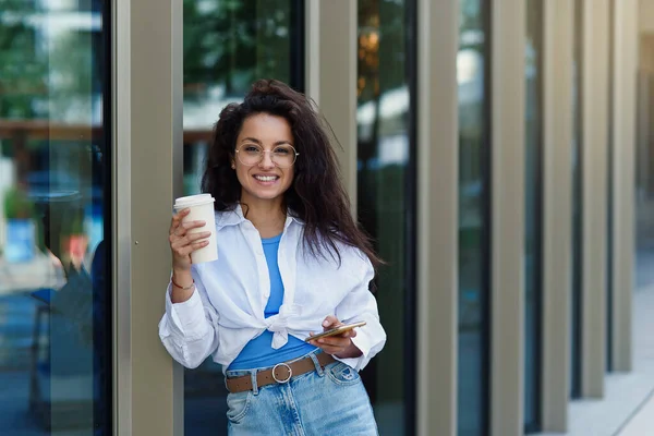 Attractive female manager with cup of tasty coffee with smartphone near business centrum. Young female company manager.