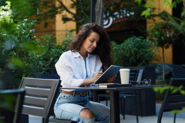 Young business woman with cup of coffee and working with tablet in cozy street cafe. Freelance working concept.