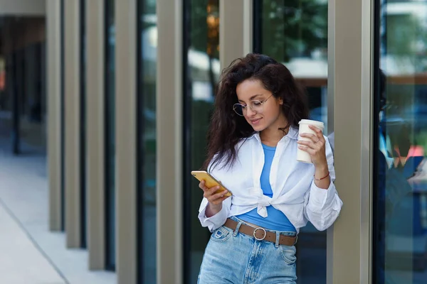 Attractive female manager with cup of tasty coffee with smartphone near business centrum. Young female company manager.