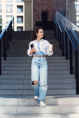Young cheerful student woman in glasses goes down the stairs with cup of coffee and notebook in her hand. Cityspace.