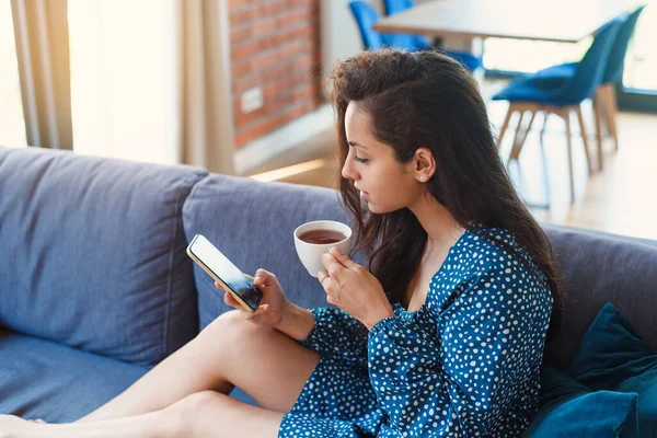 Attractive young woman chatting on smartphone and drink tea in modern living room at home. Spending leisure chatting in social network, freelancer working online