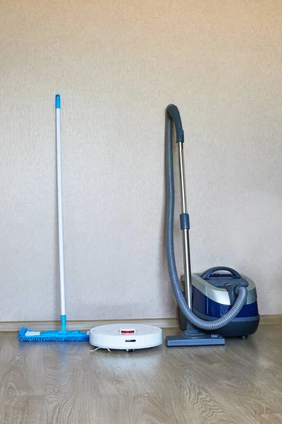 White robotic vacuum cleaner next to a conventional vacuum cleaner and a mop on laminate wood floor — Stock Photo, Image