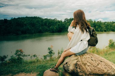 Genç Asyalı yalnız kadın yazın dağların arasında nehrin kenarında taşın başında otururken. Beyaz bir gömlek ve omuz çantası giyiyordu, sorunlarını düşünüyordu ve çok üzgündü..