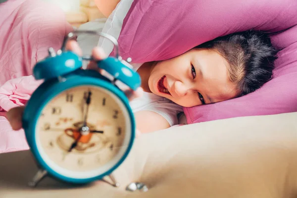 Young beautiful girl hates waking up early in the morning. Sleepy girl looking at alarm clock and trying to hide under the pillow. Students wake up early to go to school.