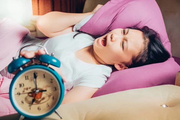 Young beautiful girl hates waking up early in the morning. Sleepy girl looking at alarm clock and trying to hide under the pillow. Students wake up early to go to school.