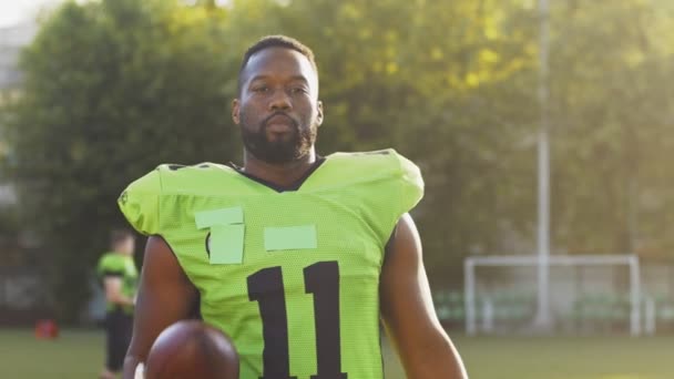 Portræt af multiraciale mandlige amerikanske fodboldspiller ser ind i kameraet og leger med bolden på grønt stadion baggrund. Sport koncept – Stock-video