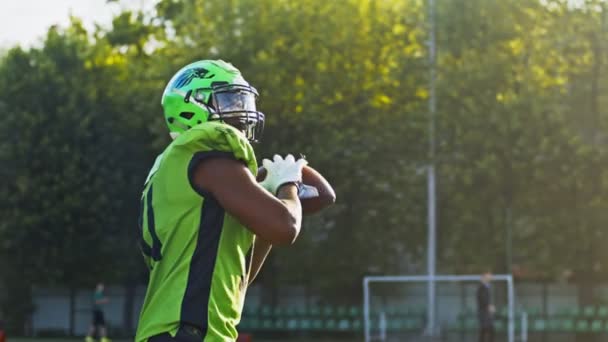 Vista lateral do jogador de futebol americano masculino em pé em seu capacete de proteção contra o estádio brilhante sob a luz do sol e jogando bola. Conceito de desporto — Vídeo de Stock
