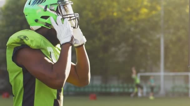 Portraitaufnahme des männlichen American-Football-Spielers, der seinen Schutzhelm gegen helles Stadionlicht im Sonnenlicht aufsetzt und in die Kamera blickt. Sportkonzept — Stockvideo