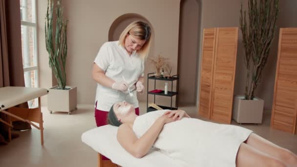 Skillful cosmetician is making healthy facial mask for client. She is standing and touching brush to female face. Young woman is lying with closed eyes and relaxing — Stock Video