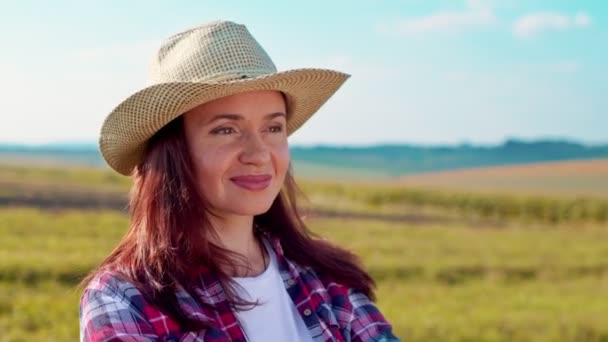 Egy mosolygó női gazda portréja, kalapban a gabonafélék között. A napfényben nő pózol munka után, mosolyog és a kamerába néz. — Stock videók