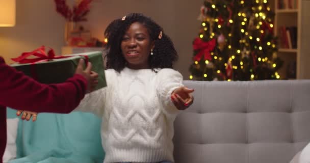 Ritratto ravvicinato di felice sorpresa afroamericana giovane mamma seduta sul divano in camera decorata con albero incandescente ricevere regalo di Natale da piccolo figlio in serata, abbracciando bambino carino. Vacanze in famiglia — Video Stock