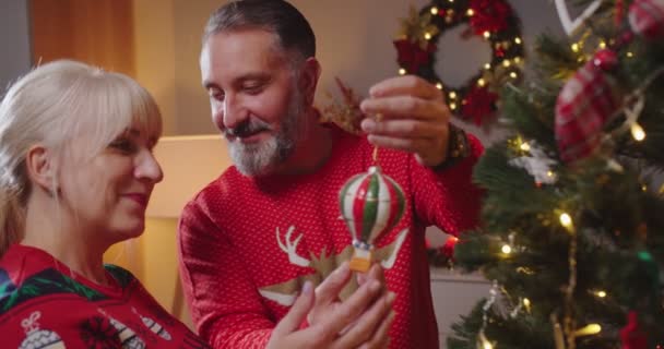Caucásico alegre pareja mayor hombre y mujer en suéteres de Navidad juntos decorando el árbol de Navidad con juguetes que se preparan para el Año Nuevo en una habitación acogedora con luces centelleantes en la noche. Concepto de primer plano — Vídeo de stock