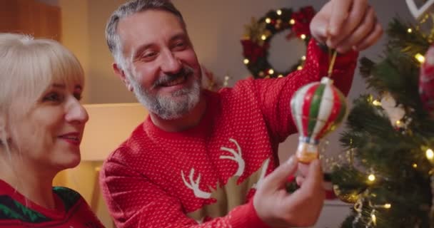 Радостная кавказская пара средних лет в рождественских свитерах украшает елку игрушками и мячами, готовясь к Новому году в уютной комнате вечером. Зимние каникулы. Закрыть — стоковое видео