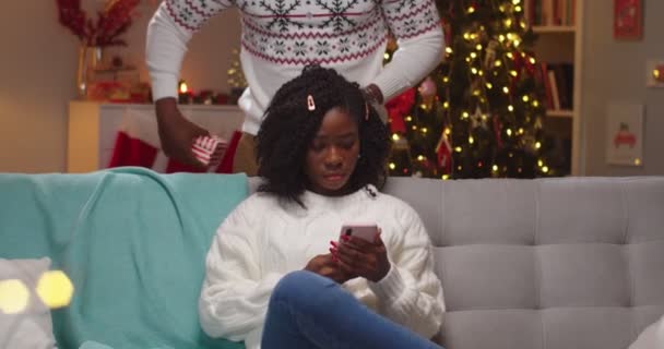 Gros plan portrait de femme afro-américaine joyeuse assis sur le canapé et tapotant sur smartphone recevant boîte cadeau de Noël de mari aimant le soir de Noël avec heureux visage surpris. Concept Nouvel An — Video