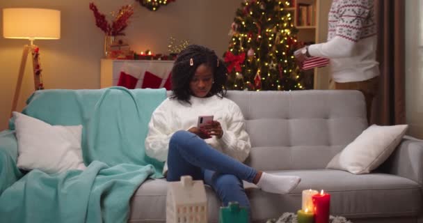 Glada afroamerikanska kvinna sitter på soffan och sms:ar på smartphone ta emot julklapp från kärleksfull make med glada förvånad ansikte på julafton. Nyårskonceptet. Glad helg — Stockvideo