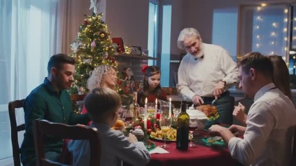 Feliz familia caucásica celebrando la Navidad, charlando en la mesa de la cena en casa. Abuelo corta pavo asado mientras todo el mundo está mirando. — Vídeos de Stock