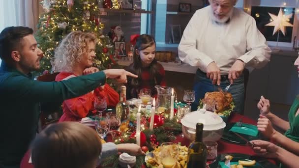 Família caucasiana feliz comemorando o Natal, conversando na mesa de jantar em casa. Avô corta peru assado enquanto todo mundo está assistindo. — Vídeo de Stock
