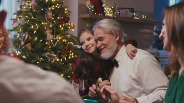 Счастливого кавказского деда, сидящего за обеденным столом со своей семьей, празднующей Рождество. Маленькая застенчивая внучка обнимает дедушку во время рождественского ужина дома. — стоковое видео