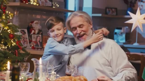 Szczęśliwego białego dziadka siedzącego przy stole z rodziną świętującą Boże Narodzenie. Mały nieśmiały wnuk daje uściski dziadkowi podczas świątecznej kolacji w domu. — Wideo stockowe