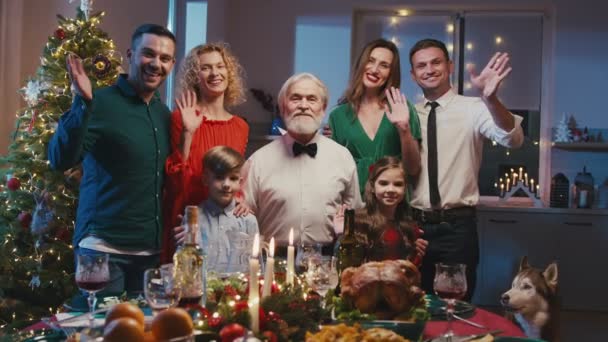 Noel yemeğinde masanın yanında duran, el sallayan ve kameraya gülümseyen mutlu aile portresi. Güzel bir aile arkadaşlarıyla ya da akrabalarıyla selfie ya da video görüşmesi yapıyor.. — Stok video