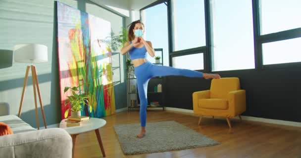 Ziemlich sportliche kaukasische Athletin in medizinischer Maske, die zu Hause trainiert und dabei starke Schwellungen aufbaut. Schöne Frau macht Fitnessübungen im Wohnzimmer. Sport-Lifestyle-Konzept — Stockvideo