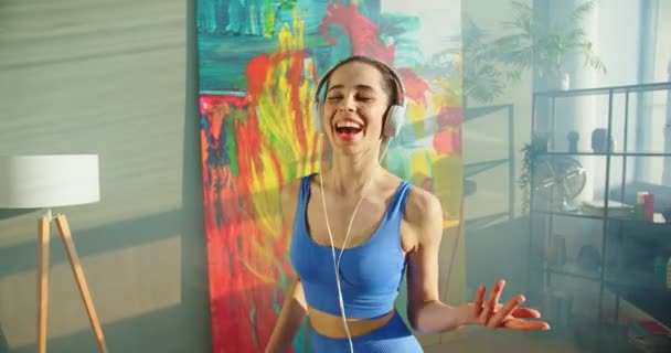 Nahaufnahme einer jungen fröhlichen kaukasischen schlanken Frau in Sportkleidung, die mit Kopfhörern Musik vom Smartphone hört und Spaß beim Singen, Tanzen und Springen zu Hause im sonnigen Raum hat Freizeitkonzept — Stockvideo