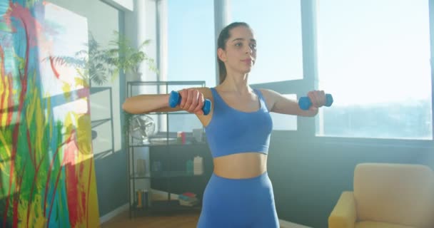 Retrato de una joven hermosa y alegre deportista caucásica haciendo ejercicios entrenando los músculos del brazo en casa usando pesas en una habitación soleada. Fitness, entrenamiento, construcción de cuerpo fuerte. Concepto de estilo de vida — Vídeos de Stock