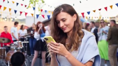 Akıllı telefonda daktilo eden genç ve güzel bir kadının portresi. Çekici kadın gülümsüyor, partideki arkadaşlarıyla iyi vakit geçiriyor. Konser, çatı partisi, bereket konsepti.
