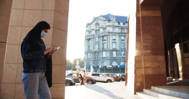 Μουσουλμάνα γυναίκα υπάλληλος στέκεται έξω από εμπορικό εταιρικό συγκρότημα φορώντας μαντίλα χιτζάμπ χρησιμοποιώντας κινητό smartphone. Χαμογελώντας ευτυχισμένη Ισλαμική κυρία γραφείο πληκτρολογώντας σε ψηφιακή gadget. Εφαρμογή αναζήτησης χαρτών. — Αρχείο Βίντεο