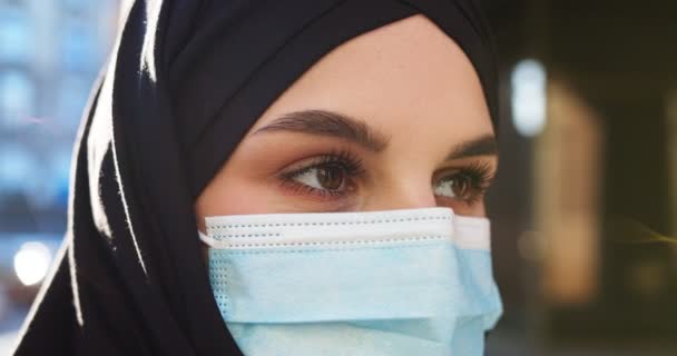 Πορτρέτο της όμορφης νεαρής Άραβας μουσουλμάνα γυναίκα σε μαύρο παραδοσιακό χιτζάμπ κοιτάζοντας κατ 'ευθείαν στην κάμερα. Στο σπίτι. Κοντινό πλάνο του θηλυκό όμορφο πρόσωπο στην ιατρική μάσκα και το Ισλάμ μαντίλα εξωτερική. Υγεία. — Αρχείο Βίντεο