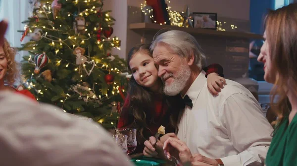 Heureux grand-père caucasien assis à la table du dîner avec sa famille célébrant Noël. Petite petite-fille timide donnant câlins à grangfather tout en ayant le dîner de Noël à la maison. — Photo