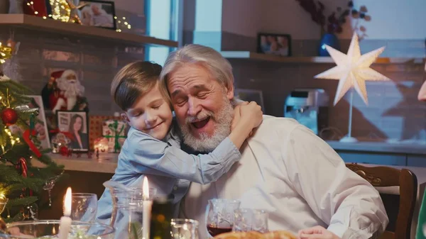 Heureux grand-père caucasien assis à la table du dîner avec sa famille célébrant Noël. Petit-fils timide donnant câlins à grangfather tout en ayant le dîner de Noël à la maison. — Photo