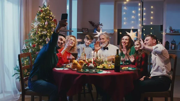 Heureuse famille caucasienne célébrant Noël, assise à la table du dîner. Belle famille souriante faisant selfie ou appel vidéo avec un smartphone à des amis ou des parents pendant le repas de famille. — Photo