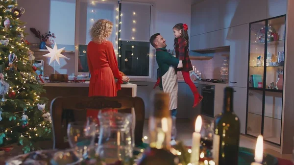 Une famille heureuse cuisine et dîne ensemble à Noël. Mère pepares nourriture et jolie petite fille court à père et ils dansent. — Photo
