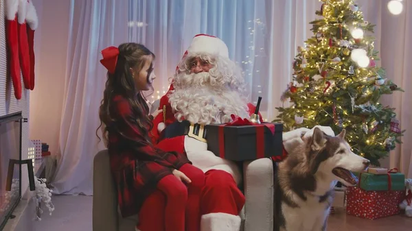 Little cute girl siedzi w pobliżu kominka na kolanach Mikołaja, szepcząc życzenie na ucho Mikołaja. Whitebroded Santa przytula ją i trzyma prezent. — Zdjęcie stockowe