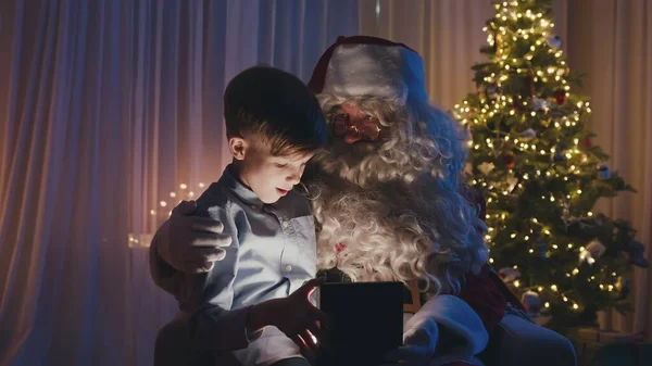 Mały, słodki chłopiec siedzący na kolanach Mikołaja, otwierający prezent przed choinką. Urocze dziecko jest szczęśliwe i zaskoczone.. — Zdjęcie stockowe