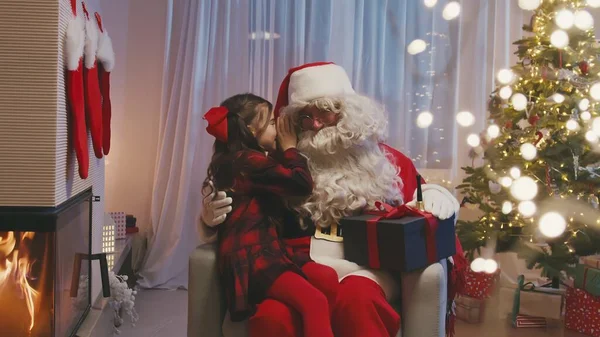 Petite fille mignonne assise près de la cheminée sur les genoux du Père Noël, chuchotant un souhait sur l'oreille du Père Noël. Le Père Noël barbu la serre dans ses bras et lui offre un cadeau. — Photo