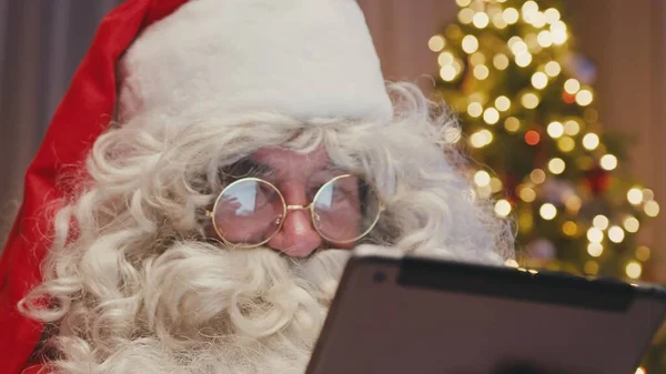 Wesoły Święty Mikołaj siedzi przed choinką wpisując na tabliczce, robiąc zakupy. Koncepcja świątecznego ducha, świąt i uroczystości. — Zdjęcie stockowe