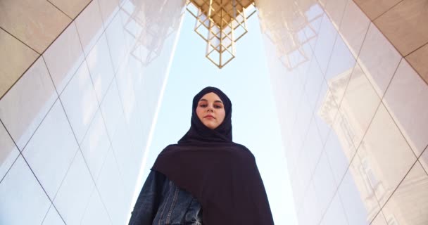 Serieuze islam dame draagt zwarte sluier op hoofd staande onder massief dak van modern gebouw. Volwassen Arabische vrouw in moslim hijab hoofddoek kijkend naar camera. Nadeel, religie, lifestyle concept. — Stockvideo